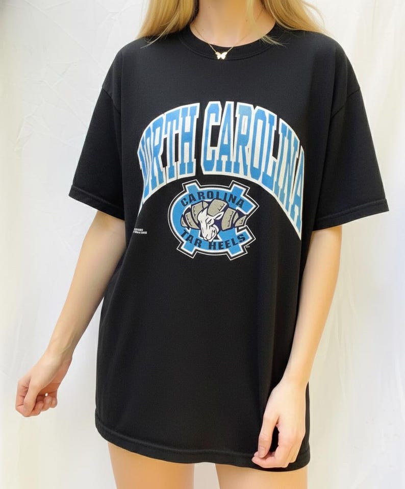 May include: Black t-shirt featuring the text "NORTH CAROLINA" in white and light blue arched lettering. Below the text is a light blue and white logo with a ram's head and the words "CAROLINA TAR HEELS".