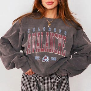 May include: A gray sweatshirt with the text "COLORADO AVALANCHE" in a distressed, maroon font. Below the text is the team logo and the year "1995". The sweatshirt has long sleeves and a crew neck.