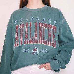 May include: A teal-colored sweatshirt with the text "COLORADO AVALANCHE" in maroon lettering. Below the text is the team logo and the text "EST. 1995". The sweatshirt has a crew neck and long sleeves.
