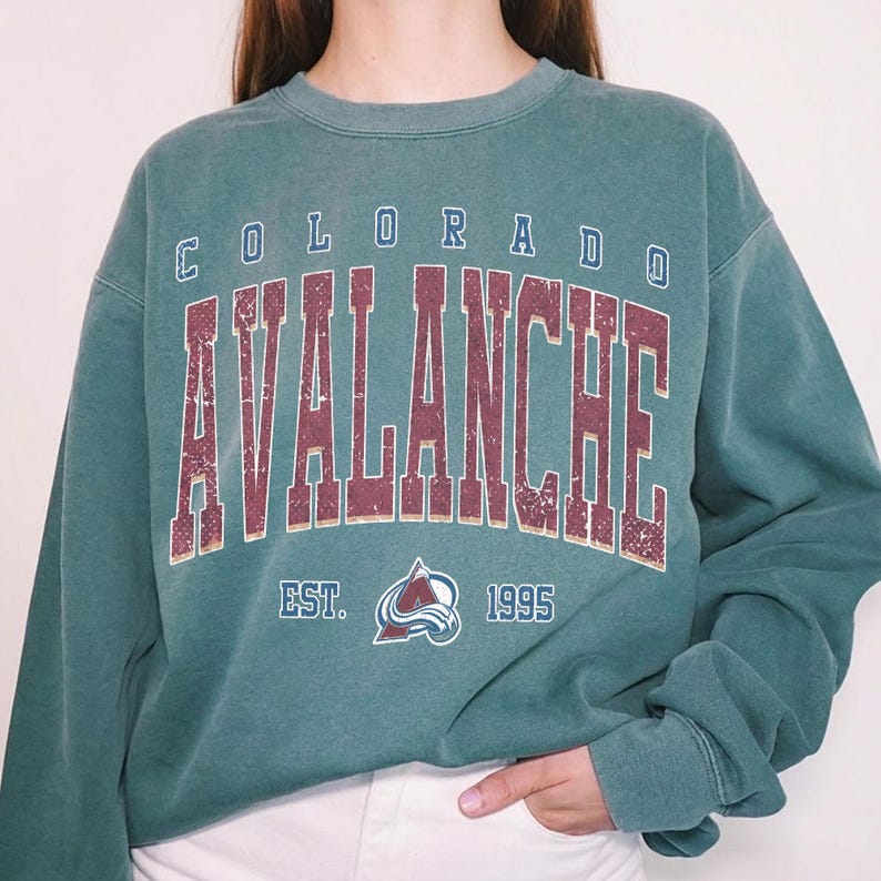 May include: A teal-colored sweatshirt with the text "COLORADO AVALANCHE" in maroon lettering. Below the text is the team logo and the text "EST. 1995". The sweatshirt has a crew neck and long sleeves.