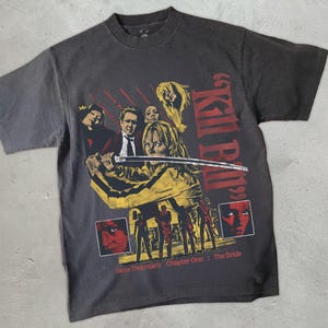 May include: Dark gray t-shirt featuring a movie poster design. The graphic includes a woman in a yellow outfit holding a sword, with the words "Kill Bill" and "Uma Thurman's Chapter One: The Bride" in red.