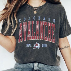 May include: Dark gray t-shirt featuring the text "COLORADO AVALANCHE" in a distressed, vintage style. The shirt includes the team logo and the year "1995". The shirt is a crew neck and has short sleeves.