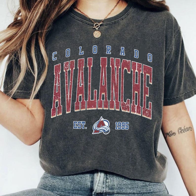 May include: Dark gray t-shirt featuring the text "COLORADO AVALANCHE" in a distressed, vintage style. The shirt includes the team logo and the year "1995". The shirt is a crew neck and has short sleeves.