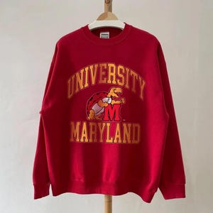 May include: A red crewneck sweatshirt featuring the University of Maryland logo. The logo includes the words "UNIVERSITY" and "MARYLAND" in gold lettering, along with a cartoon turtle mascot. The sweatshirt is hanging on a wooden hanger.