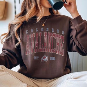 May include: A brown sweatshirt with the text "COLORADO AVALANCHE" in a distressed, vintage style. The sweatshirt also features the team's logo and the year "1995". The sweatshirt is a comfortable, casual style.