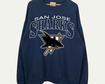 May include: Navy blue sweatshirt featuring the San Jose Sharks logo. The logo includes the team name in white lettering above a black shark with a hockey stick inside a white triangle. The sweatshirt is hanging on a wooden hanger.
