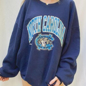 May include: Navy blue sweatshirt with the words "North Carolina" in light blue arched across the top. Below the text is a logo with a ram and the words "Tar Heels". The sweatshirt has long sleeves and a crew neck.