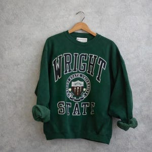 May include: A green sweatshirt with the words "WRIGHT STATE UNIVERSITY" in white block letters. The university's emblem is in the center. The sleeves are rolled up. The sweatshirt is hanging on a wooden hanger.