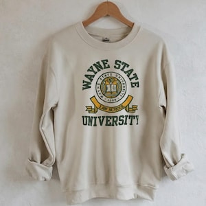May include: A beige sweatshirt with the Wayne State University Law School emblem in green and yellow. The emblem features a circular design with the university name, a book, and the year 1869. The sweatshirt is hanging on a wooden hanger.