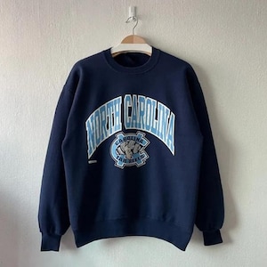 May include: Navy blue sweatshirt with the words "NORTH CAROLINA" in white arched lettering. Below the text is a light blue emblem with the words "TAR HEELS". The sweatshirt is hanging on a wooden hanger.