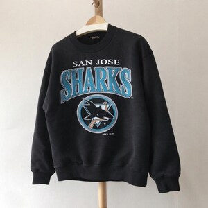 Peut inclure: Sweat-shirt noir des Sharks de San José avec le nom et le logo de l'équipe en bleu sarcelle et blanc. Le logo représente un requin avec une crosse de hockey et le texte "SAN JOSE SHARKS" au-dessus.