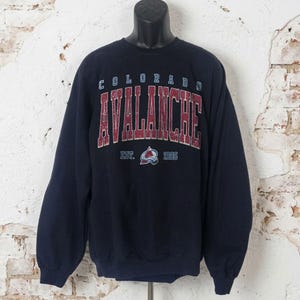May include: Navy blue sweatshirt featuring the text "COLORADO AVALANCHE" in maroon and white lettering. The sweatshirt also displays the team logo and the text "EST. 1995". The long-sleeved top is displayed on a black mannequin.