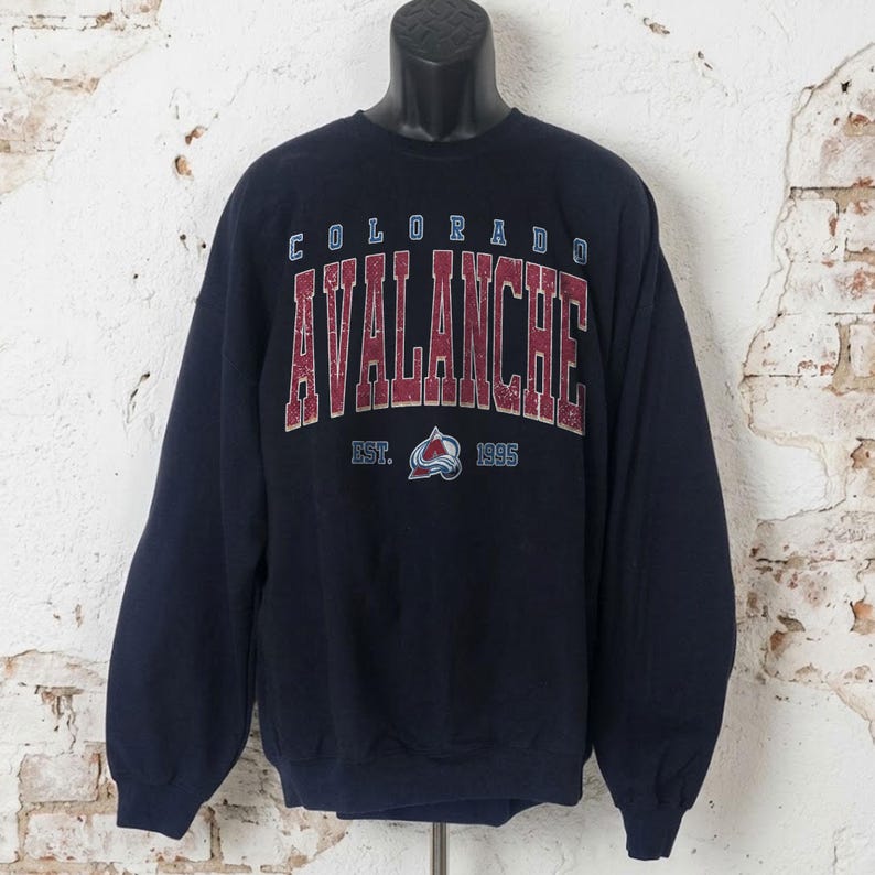 May include: Navy blue sweatshirt featuring the text "COLORADO AVALANCHE" in maroon and white lettering. The sweatshirt also displays the team logo and the text "EST. 1995". The long-sleeved top is displayed on a black mannequin.