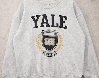 May include: A light gray sweatshirt with the word "YALE" in large black letters across the chest. Below the word is a crest with the Latin phrase "LUX ET VERITAS" and a gold wreath. The sweatshirt is a crew neck style.