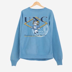 May include: Light blue sweatshirt with the letters "UNC" in navy blue across the chest. A cartoon ram mascot is centered below the letters, within a yellow and navy blue diamond. The words "University of North Carolina" are printed below the mascot.