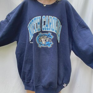 May include: Navy blue sweatshirt with the words "North Carolina" in light blue block letters. Below the text is a circular emblem with a ram and the words "Carolina Tar Heels". The sweatshirt is oversized.