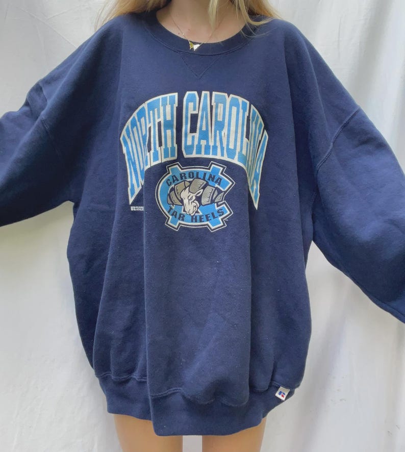 May include: Navy blue sweatshirt with the words "North Carolina" in light blue block letters. Below the text is a circular emblem with a ram and the words "Carolina Tar Heels". The sweatshirt is oversized.