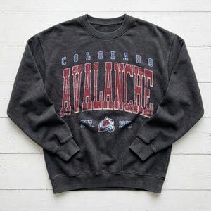 May include: A charcoal gray sweatshirt with the words "COLORADO AVALANCHE" in a distressed, vintage style. The text is in red and blue, with the team logo below. The sweatshirt has a crew neck and long sleeves.