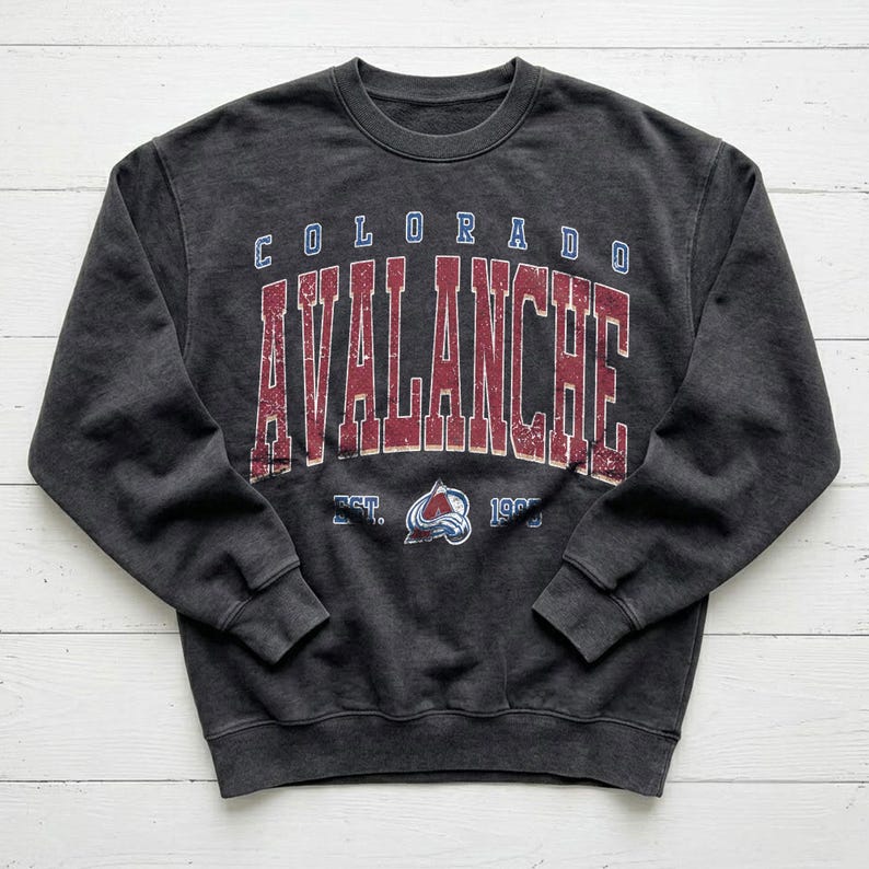 May include: A charcoal gray sweatshirt with the words "COLORADO AVALANCHE" in a distressed, vintage style. The text is in red and blue, with the team logo below. The sweatshirt has a crew neck and long sleeves.