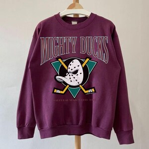 May include: A maroon sweatshirt featuring the text "MIGHTY DUCKS" in white arched lettering. Below the text is a teal triangle with a hockey mask and crossed hockey sticks. The text "SEASONAL SEASON 1992-1993" is below the triangle.