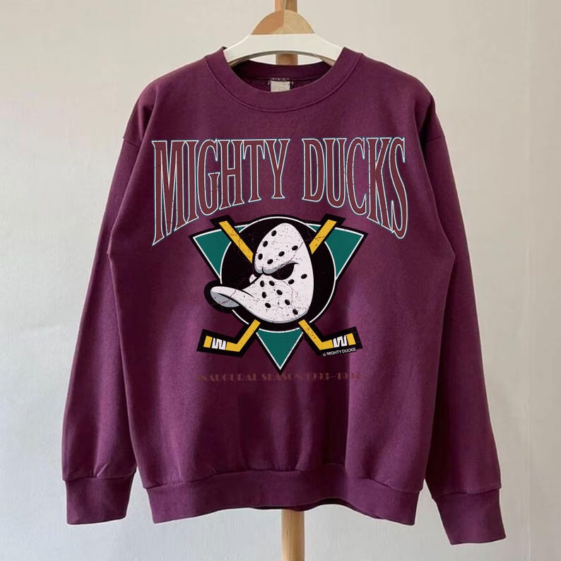 May include: A maroon sweatshirt featuring the text "MIGHTY DUCKS" in white arched lettering. Below the text is a teal triangle with a hockey mask and crossed hockey sticks. The text "SEASONAL SEASON 1992-1993" is below the triangle.