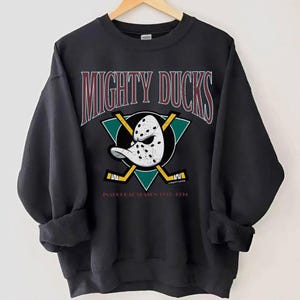May include: Black sweatshirt featuring the text "MIGHTY DUCKS" in maroon and white. The design includes a hockey mask, crossed hockey sticks, and a teal triangle. The text "INAUGURAL SEASON 1993-1994" is also visible.