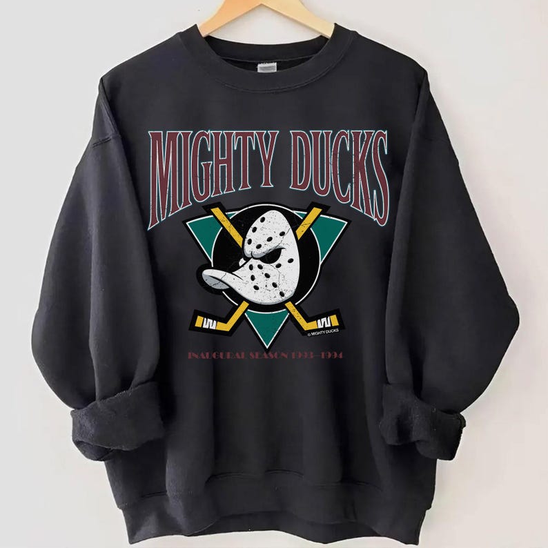 May include: Black sweatshirt featuring the text "MIGHTY DUCKS" in maroon and white. The design includes a hockey mask, crossed hockey sticks, and a teal triangle. The text "INAUGURAL SEASON 1993-1994" is also visible.