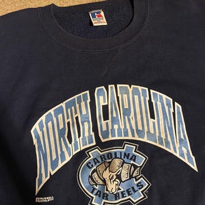 May include: Navy blue sweatshirt with the words "NORTH CAROLINA" in white and light blue arched across the chest. Below the text is a light blue and navy logo with a ram and the words "CAROLINA TAR HEELS". The sweatshirt has a crew neck.