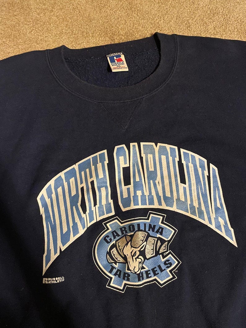 May include: Navy blue sweatshirt with the words "NORTH CAROLINA" in white and light blue arched across the chest. Below the text is a light blue and navy logo with a ram and the words "CAROLINA TAR HEELS". The sweatshirt has a crew neck.