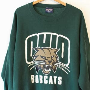 May include: A dark green sweatshirt with the word "OHIO" in white block letters above a brown and tan bobcat graphic. Below the graphic, the word "BOBCATS" is printed in white block letters. The sweatshirt is hanging on a wooden hanger.