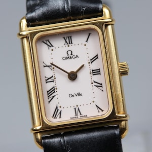 May include: A rectangular gold-tone Omega De Ville wristwatch featuring a white dial with black Roman numeral hour markers, a black leather strap, and the text Omega, De Ville, and Swiss Made printed on the face with gold-tone hands.