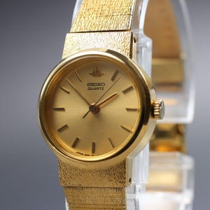 May include: A gold-tone Seiko Quartz wristwatch featuring a round gold dial with thin gold hour markers and hands. The watch has a textured gold-tone metal link bracelet and a crown on the right side. The Seiko logo is printed on the dial.