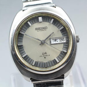May include: A silver-toned Seiko automatic wristwatch featuring a round face with a light gray center, a dark gray outer ring, and silver hour markers. The dial displays the text Seiko, Automatic, LM, 23 Jewels, and a day-date window showing 26.