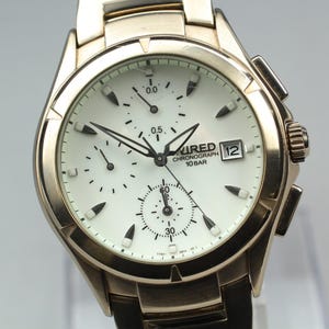 May include: A rose gold-tone chronograph wristwatch with a white dial, featuring three sub-dials, a date window at the three o'clock position, and the text WIRED CHRONOGRAPH 10 BAR printed on the face. The watch has a metallic link bracelet.