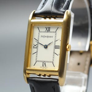 Könnte beinhalten: Eine rechteckige Armbanduhr mit goldfarbenem Gehäuse und cremefarbenem Zifferblatt. Das Zifferblatt zeigt schwarze römische Ziffern, einen zentralen rechteckigen Rahmen und den Schriftzug Yves Saint Laurent. Sie hat ein schwarzes Lederarmband.