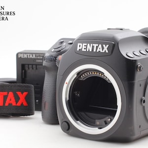 Puede incluir: Una cámara digital Pentax negra con montura de objetivo y el nombre de la marca en blanco. También se ven un cargador de batería negro y una correa con el logotipo de Pentax en rojo. La cámara está sobre un fondo blanco.