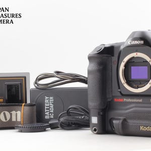 Peut inclure: Un boîtier d'appareil photo numérique noir Canon EOS-1n Kodak Professional DCS 520 avec monture d'objectif, un adaptateur secteur avec câble, une sangle noire avec le logo Canon et un bouchon d'objectif. L'appareil porte la marque Kodak.