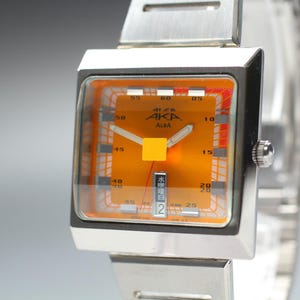 May include: A square-faced wristwatch with a silver-tone metal band and case. The dial is bright orange with a white grid pattern, featuring the text "AKA" and "ALBA" in black. It includes silver hands, a date window, and a small yellow square.