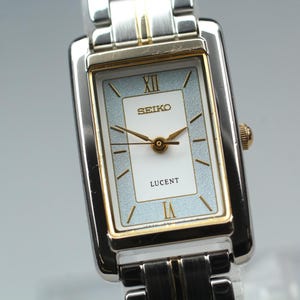 Puede incluir: Un reloj de pulsera rectangular Seiko Lucent con una correa de eslabones de acero inoxidable en dos tonos, plata y oro. La esfera tiene un centro blanco, un borde texturizado azul claro, manecillas, marcadores y números romanos dorados.