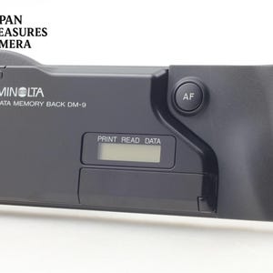 May include: A black Minolta Data Memory Back DM-9 camera accessory featuring a digital display screen with the text PRINT READ DATA, a circular AF button, and a contoured grip section on the right side, designed for specific camera models.