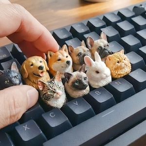 May include: A collection of animal-themed keycaps, including dogs, cats, and a rabbit, are displayed on a black keyboard. The keycaps are intricately detailed and add a unique touch to the keyboard. The hand is holding one of the keycaps.