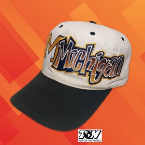 Könnte beinhalten: Eine Baseballkappe mit weißer Krone und schwarzem Schirm. Auf der Vorderseite ist das Wort Michigan in großen, fetten Buchstaben mit einer gold- und marineblauen Farbverlaufsfüllung und weißer Umrandung aufgestickt. Die Kappe hat dunkle Ösen.