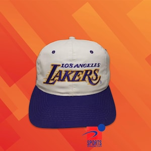 May include: A white baseball cap with a purple brim featuring the Los Angeles Lakers logo embroidered in purple and yellow on the front. The cap has a structured crown with eyelets and a button on top, displaying the Sports Specialties logo.
