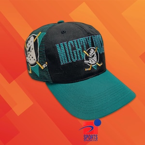 以下が含まれることがあります： フロントパネルにティールカラーでMIGHTY DUCKSと刺繍された、黒とティールの野球帽。キャップの前面と側面には、白いアヒルのマスクのロゴと交差したホッケースティックが刺繍されており、ティールカラーのつばとメッシュの背面を備えています。
