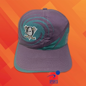 Op de afbeelding: Een paarse honkbalpet met een geborduurd blauwgroen wervelontwerp en een ijshockeymaskerlogo in het midden. De pet heeft een blauwgroene rand aan de klep en een gestructureerde kroon. Het logo toont een eendvormig keepersmasker met hockeysticks.