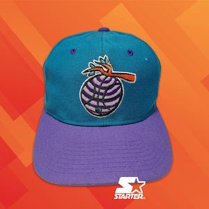Pode incluir: Um boné de beisebol azul-petróleo com aba roxa e um botão roxo no topo. A frente apresenta um gráfico bordado de uma bola de basquete com listras roxas e brancas e um design em forma de galho laranja e azul-petróleo no topo da bola.