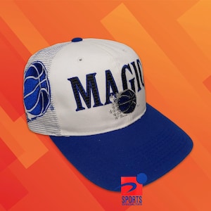 Op de afbeelding: Een wit en blauwe honkbalpet met het woord MAGIC geborduurd in blauw met zwarte omlijning. Het zijpaneel toont een blauwe basketbalafbeelding en een klein basketbalicoon is in de tekst verwerkt. De klep is effen blauw van kleur.