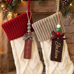 May include: Two cream-coloured cable knit stockings with ribbed cuffs. The left stocking has a red cuff and a tag reading Peace, Love, and Joy. The right stocking has a tan cuff and a tag reading Jackson. Both feature wooden bead accents.