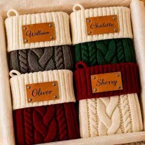 May include: Four cable-knit Christmas stockings arranged in a box. Each stocking features a cream-colored cuff with a personalized tan leather tag riveted to the front. The names William, Chalette, Oliver, and Sherry are engraved on the tags.