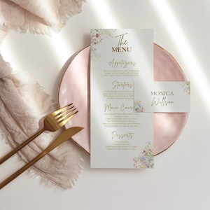 May include: A wedding menu and place card set on a pink plate, with gold cutlery and a soft, beige fabric. The menu features floral accents and gold text, including "The Menu" and the name "Monica Willson".